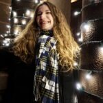 Headshot of Abigail McGovern, a white woman with long wavy brown hair. She is wearing a long black winter coat, a brown plaid skirt, and a scarf in blue, white, and yellow. She stands in between two concrete pillars, which are strung with white holiday light.