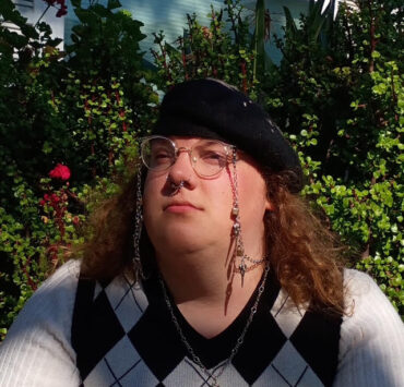 Carson Sandell, a white transfeminine person, sits in front of a bush wearing glasses, a black beret, and long sleeve sweater vest.