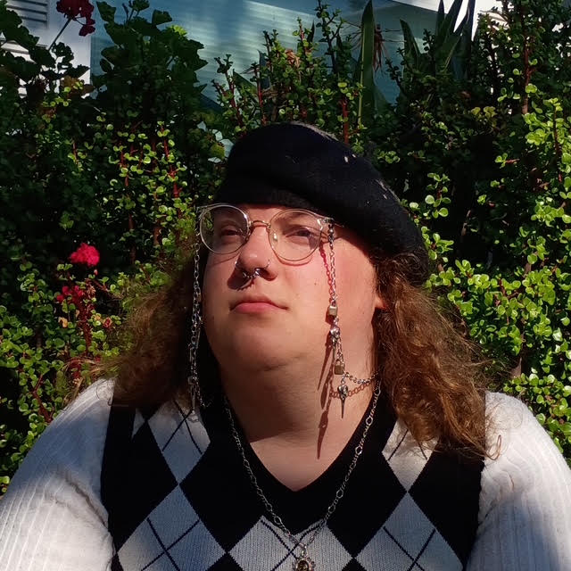 Carson Sandell, a white transfeminine person, sits in front of a bush wearing glasses, a black beret, and long sleeve sweater vest.