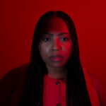 A woman with long braided hair faces the camera in a close-up portrait, wearing a red top against a vivid red background. Dramatic red lighting casts deep shadows across her face and clothing.