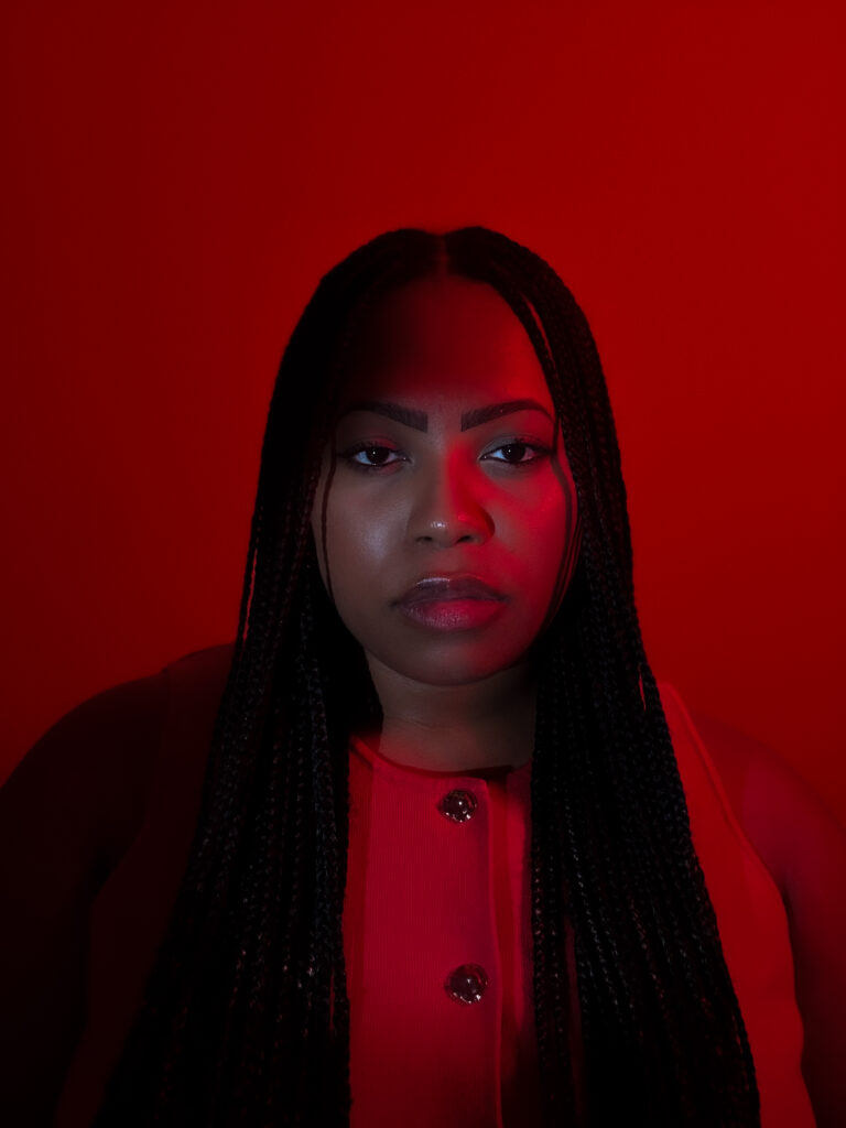 A woman with long braided hair faces the camera in a close-up portrait, wearing a red top against a vivid red background. Dramatic red lighting casts deep shadows across her face and clothing.