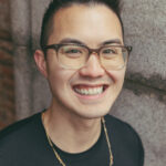 Joshua Nguyen, with a black fohawk hairstyle, smiling at the camera while wearing a black shirt with a thin, gold chain.