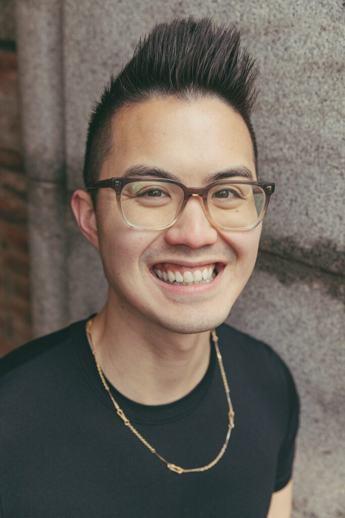 Joshua Nguyen, with a black fohawk hairstyle, smiling at the camera while wearing a black shirt with a thin, gold chain.