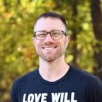 A headshot of Darren C. Demaree smiling in a park during fall with a black t-shirt on that reads "Love Will".