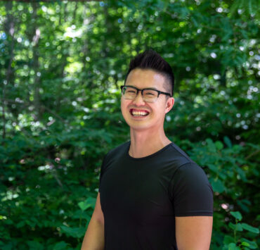 Joshua Nguyen, wearing a black shirt and glasses, smiles at the camera while standing in front of green foliage.
