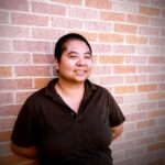 An Asian American person wearing a dark shirt stands in front of a brick wall and smiles.