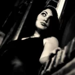 A black and white photo of a non-binary person with dark hair and dark eyes, looking up, framed by a brick wall in the background and the night sky.