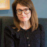 A white woman in a black dress and glasses looking at the camera.