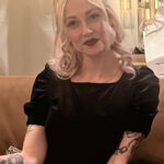 Close up photo of a blonde haired woman with tattoos in a black dress.