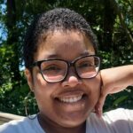 An outdoor headshot of Sienna Morgan, a woman with short curly hair, wearing black glasses and a white shirt, smiling.
