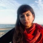 A headshot of Adriana Rambay Fernandez with long brown hair, dark eyes, and a solemn expression, wearing hoop earrings and a red wrap, set against the backdrop of blue sky, clouds, sand and ocean.