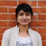 A photograph of a woman with dark hair wearing a grey jacket standing next to a brick wall.