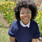 A black person sitting in front of a field of tulips, smiling widely. They have an afro and wear mint-colored glasses with a navy sweater and gray jeans.