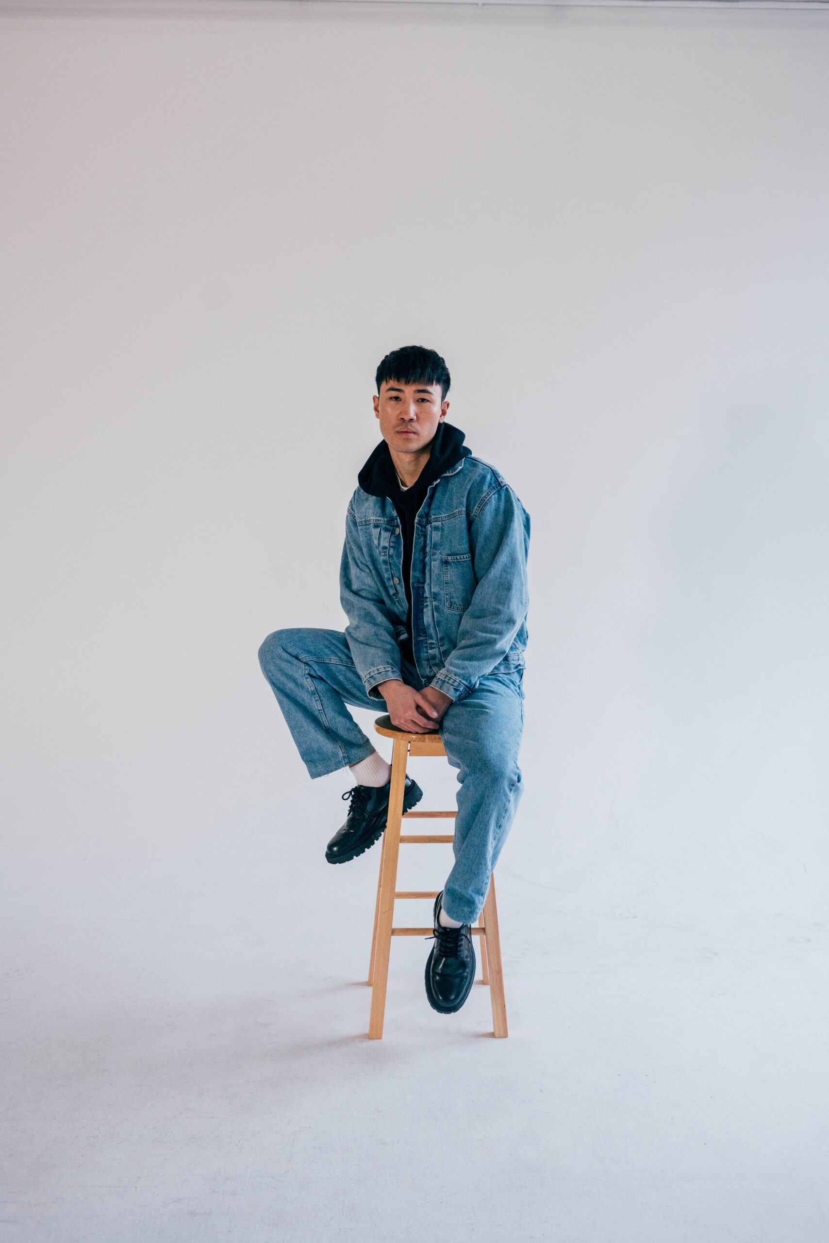 An Asian man wearing blue jeans and a blue denim jacket over a black hooded sweatshirt, posing on a wood stool against a white background.