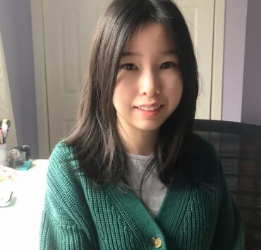 Heather is sitting on a black desk chair in a room with lavender walls. In the background is a closet with white doors, a white desk, a gray curtain, and a cup of pens.