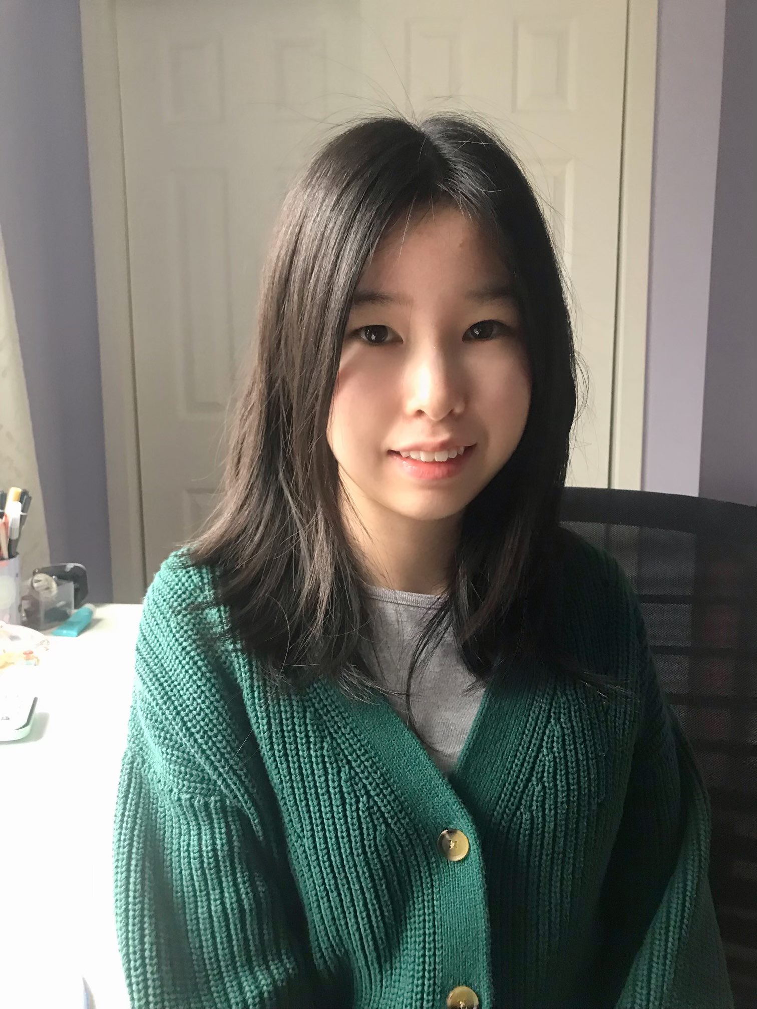 Heather is sitting on a black desk chair in a room with lavender walls. In the background is a closet with white doors, a white desk, a gray curtain, and a cup of pens.