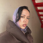 Photo of Angel Leyba, a non-binary person wearing a lavender scarf tied over their head into a bow under their chin and a dark brown puffer coat.