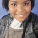 a brown skinned woman with chin length hair stands in front of an indigo colored wall. She is framed in photo from the chest up, smiling for a selfie.