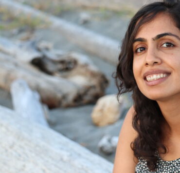 Mahreen, a brown woman with wavy shoulder-length hair, is smiling from one side of the frame, wearing a black-and-white floral top. Some grey driftwood in the blurry background can be made out.