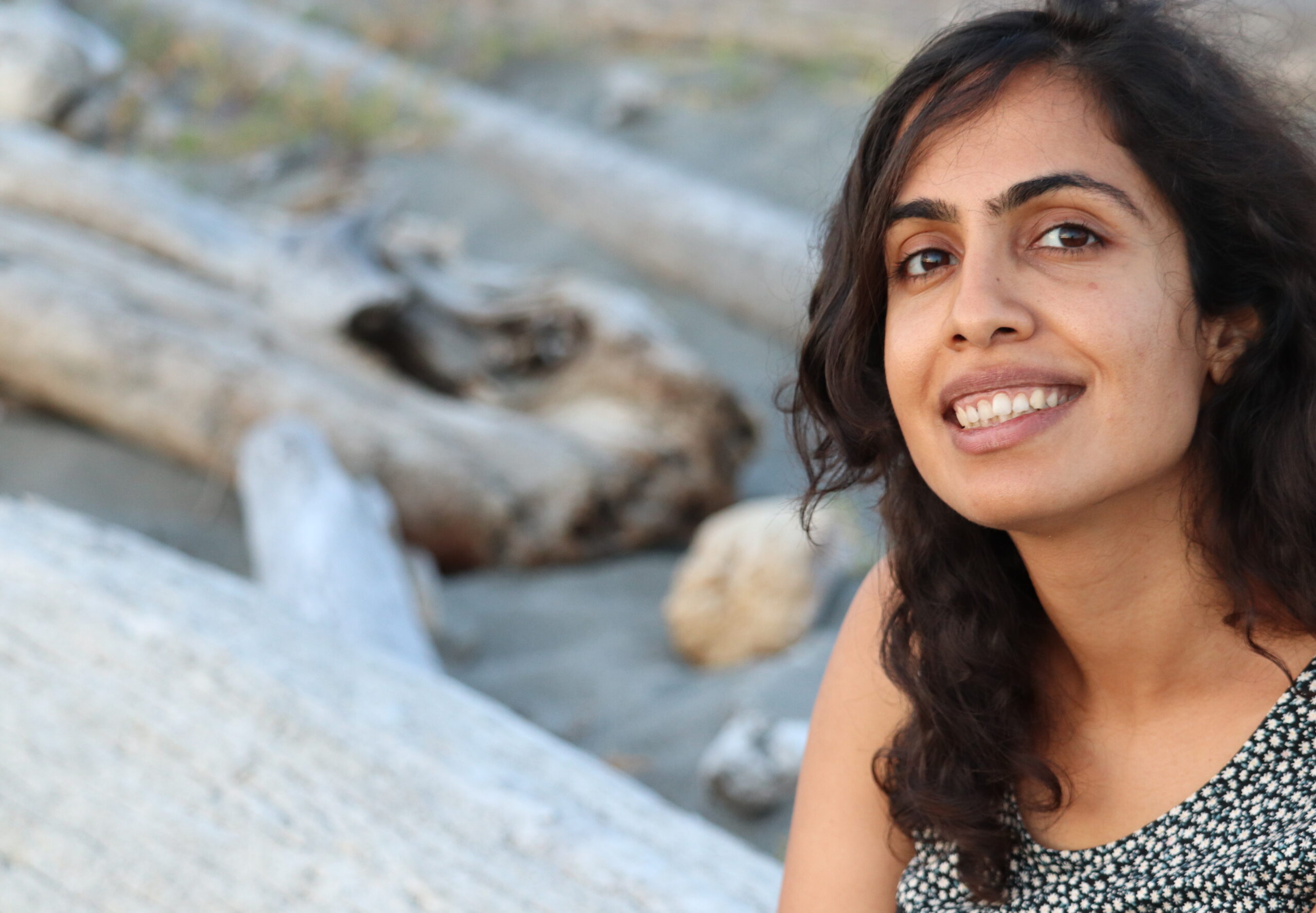 Mahreen, a brown woman with wavy shoulder-length hair, is smiling from one side of the frame, wearing a black-and-white floral top. Some grey driftwood in the blurry background can be made out.