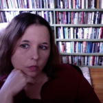 Photo of a white woman with freckles and brown hair in front of a floor to ceiling bookshelf.