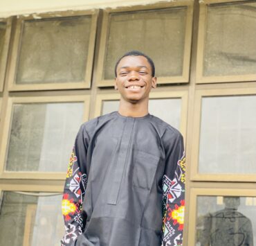 A young man smiling confidently, wearing a black traditional outfit with colorful patterned sleeves, standing in front of a large window with a bright and joyful expression.