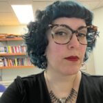 Femme-presenting person with navy curly hair and large-framed glasses sits in front of a pale wall with a bookshelf.