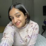 Photo of a south asian woman, Maya, in her room smiling . She is wearing a light pink turtleneck with purple flowers and gold hoop earrings.