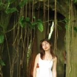 Photo of a young Asian woman with copper hair in a white dress standing under the arch of a large tree. She is looking up at the plant roots and leaves hanging above her head.