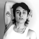 A black-and-white photo of Binx, a brunette, looking into the camera, one arm resting over their head. Photo credit to to Mary Stathos, 2023.