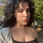 Headshot of woman with black curly hair, wearing a black dress and a white cardigan in front of trees.