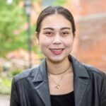 A photo of a biracial Asian Canadian femme standing in front of a blurred-out brick building with trees, a lampost, and a walkway. Their brunette hair is tied back into a bun and they are wearing a black faux leather jacket over a black top with a gold chain and a gold necklace with a "K" pendant. They are smiling at the camera.