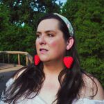 Evelyn stands on a bridge in a public park. She is wearing a green headband and large cloth earrings shaped like strawberries. She is looking off to the left of the camera.