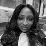 A person with long, voluminous curls takes a black-and-white selfie on a train. They wear a light-colored puffer jacket and have nose jewelry. Reflections from the train window and blurred motion of the outside world are visible in the background.