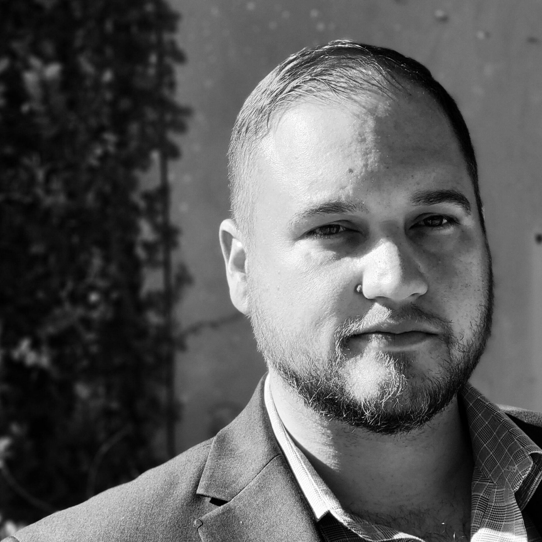 Black-and-white headshot of Clayton Bradshaw-Mittal with a short beard and short hair from the shoulders up smiling in a suit with no tie.