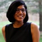 Indian woman with short dark hair, wearing a black sleeveless dress and purple glasses, smiling into the camera.