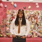 Dr. Kelsey L. Smoot (Kelz) is a Black transmasculine person with locs, wearing a maroon hat, a white button-down shirt with a dark orange tie. He is smiling, with his hands tucked into his pockets, standing in front of a multicolored art installation with dangling fixtures above his head.