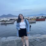 Livia is a white woman with dark brown hair. She is wearing comfortable walking clothes and is standing in front of fishing boats, a small rock wall, and Mount Vesuvius.