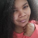 Portrait-oriented selfie of Ashley Elizabeth smirking and wearing a coral pink shirt in 3/4th profile, her long, coiled, dark brown hair over a shoulder and taking up the majority of the background, Her chin rests on her hand.