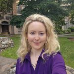 A portrait photo of Ana Mourant, a young woman, smiling, with light skin and blond hair, wearing a purple dress, outside with trees in the background.