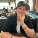 A white person in a black button-down shirt and black Oakland A's baseball cap, wearing rings on both hands, sits at a table and leans their chin on their right hand. They smile softly at the camera.
