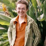 Cavar, a genderless white person with short, dark-blonde hair, glasses, and several visible facial piercings, smiles at the camera. They are wearing an orange shirt beneath a green, black, and white patterned jacket, and are standing in a thicket of deep green leaves.