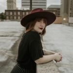 A white woman looks over her right shoulder and into the camera while standing on an empty parking garage rooftop. Her hands are clasped together as she rests on a ledge in front of her. She has short red hair with bangs. She wears a maroon wide brim hat, large silver hoop earrings, deep red lipstick, a short sleeve black shirt, and black trousers with thin white stripes.