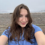 A white woman wearing a blue denim dress smiles at the camera in a "selfie" format. She has brown hair and blue eyes, while a hazy sky and a body of water appear behind her.