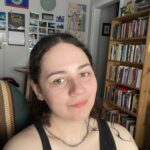 white-skinned, dark-haired person smiling. They are wearing a black tank top and silver chain necklace. In the background is a bookshelf.