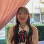 A close-up of an Asian woman with long brown hair and front bangs smiling at the camera. She is wearing a light tan cardigan and a cream-colored collar shirt with a navy blue and red ribbon tied in the front. An empty street with two parked cars is behind her and she is standing in front of a pink curtain and green hedge.