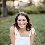 A young white woman with wavy brown hair sits on the grass, smiling at the camera. She is wearing a white sleeveless top with tie straps and layered gold necklaces, including one with a pink butterfly charm. The background is softly blurred with green trees and warm sunlight.