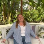 A white woman with blonde hair and clear glasses sitting on a couch outside with several trees and plants in the background. She is wearing jeans and a blue-and-white striped button down shirt.
