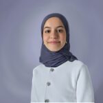 Headshot of brown woman of Middle Eastern/North African descent against a bluish-lavender background. She wears a greyish navy hijab (headscarf), silver earrings, and a white jacket with silver buttons.