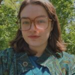 A picture of a light-skinned person with shoulder-length brown hair and large glasses sitting in a park wearing a patterned turquoise outfit and seashell necklace.